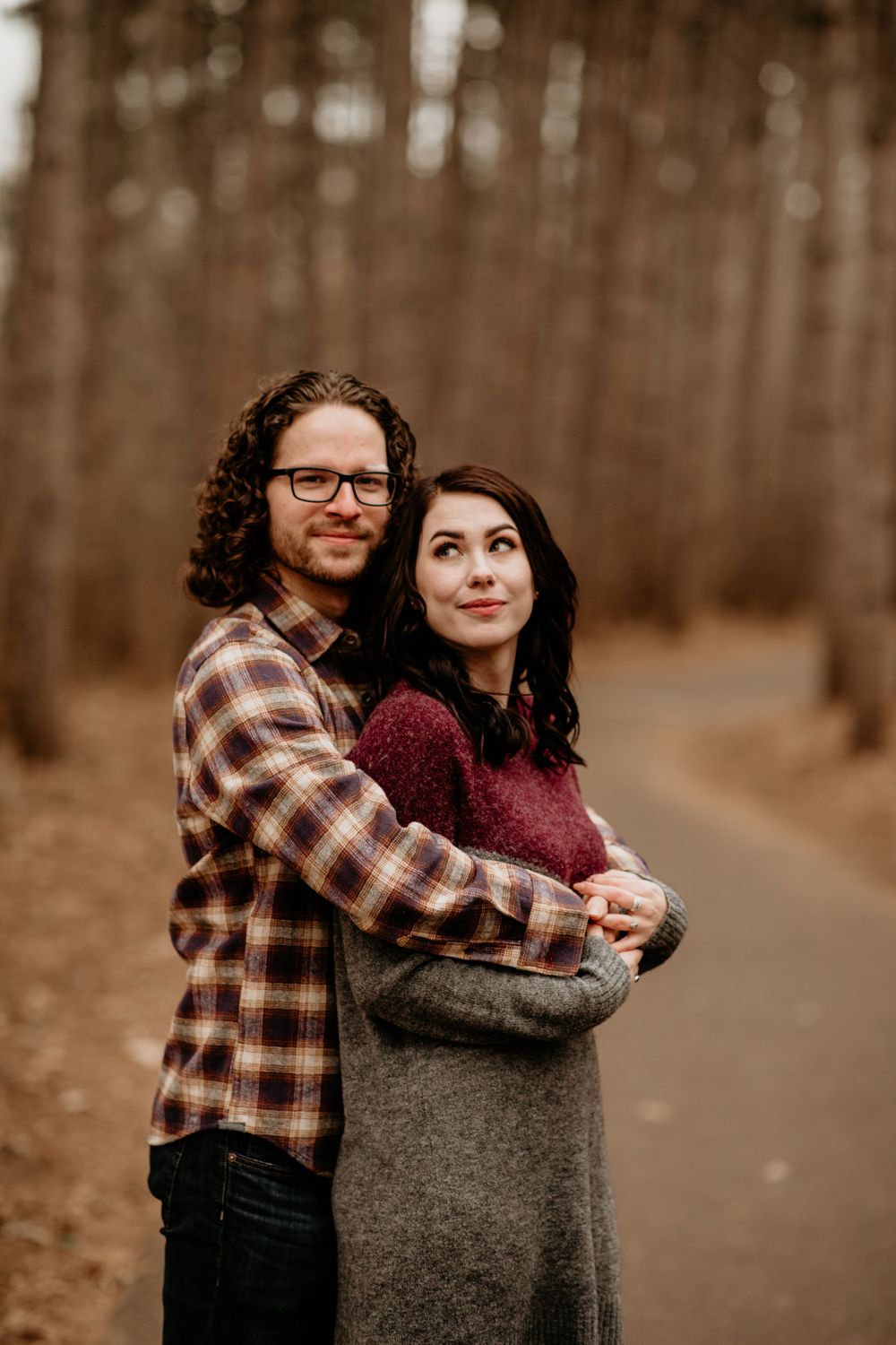 Couple posing on an autumn forest path, man in plaid shirt hugging woman in gray sweater from behind.