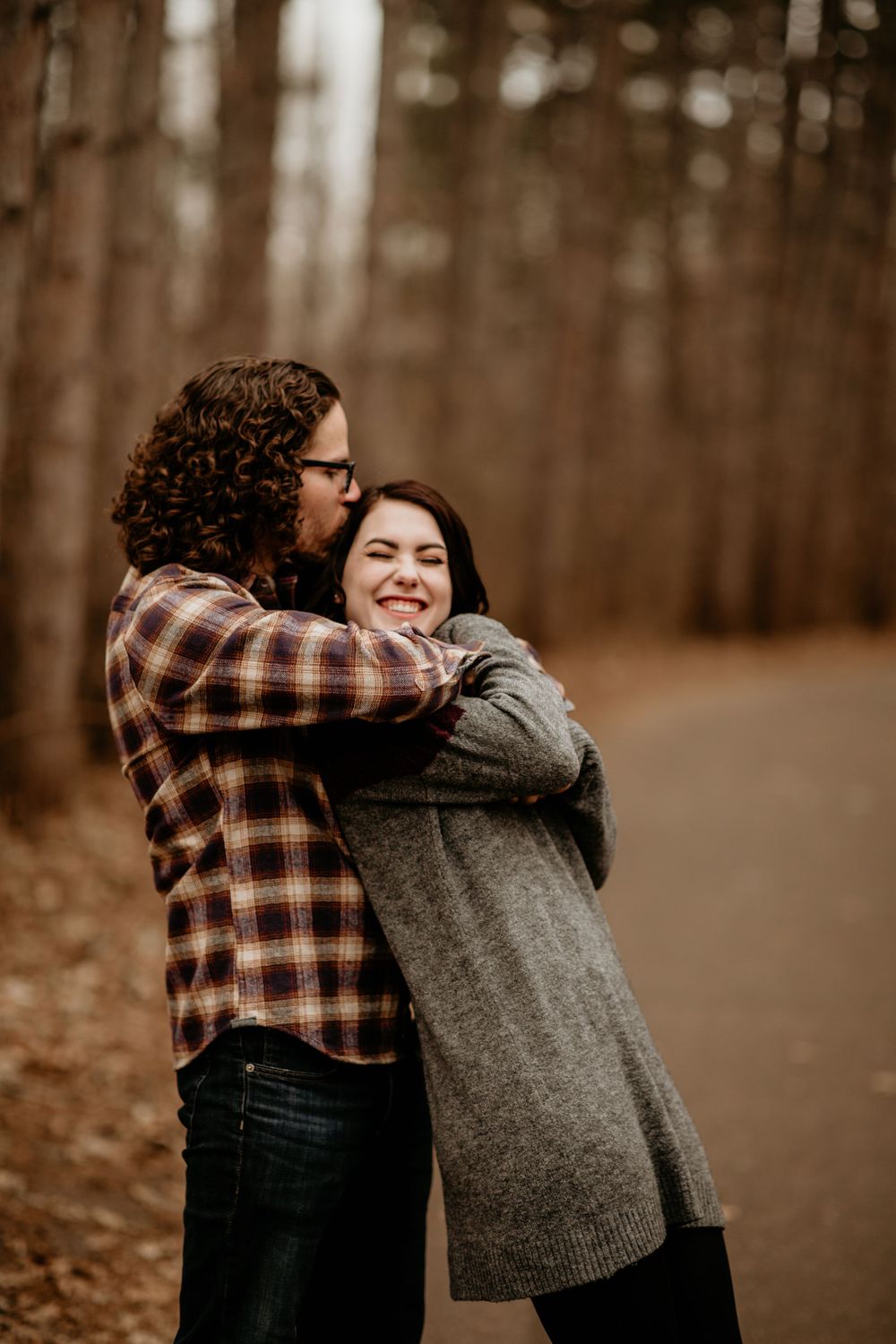 Man in plaid shirt hugging and kissing laughing woman in gray sweater on a wooded forest path.