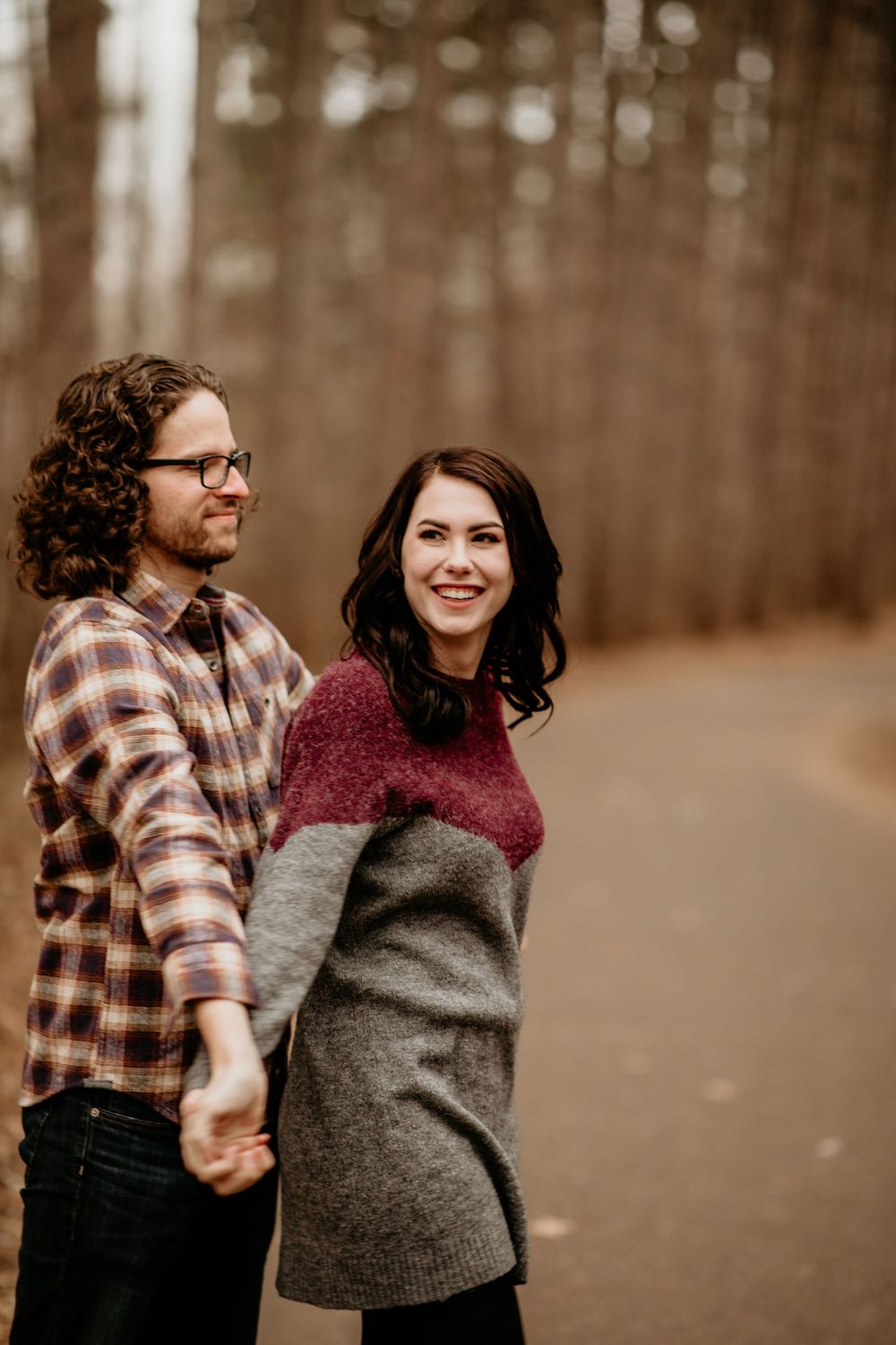 Couple holding hands on a forest path in fall, woman in colorblock sweater smiling over her shoulder.
