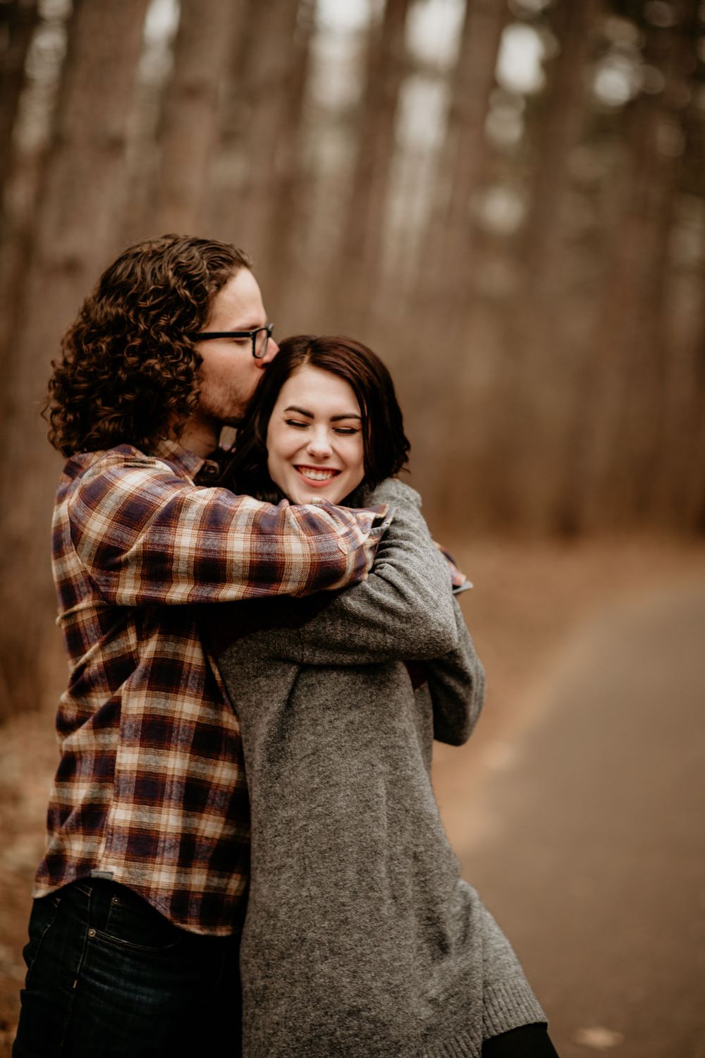 Man in plaid shirt kissing smiling woman on cheek while embracing her on an autumn forest path.