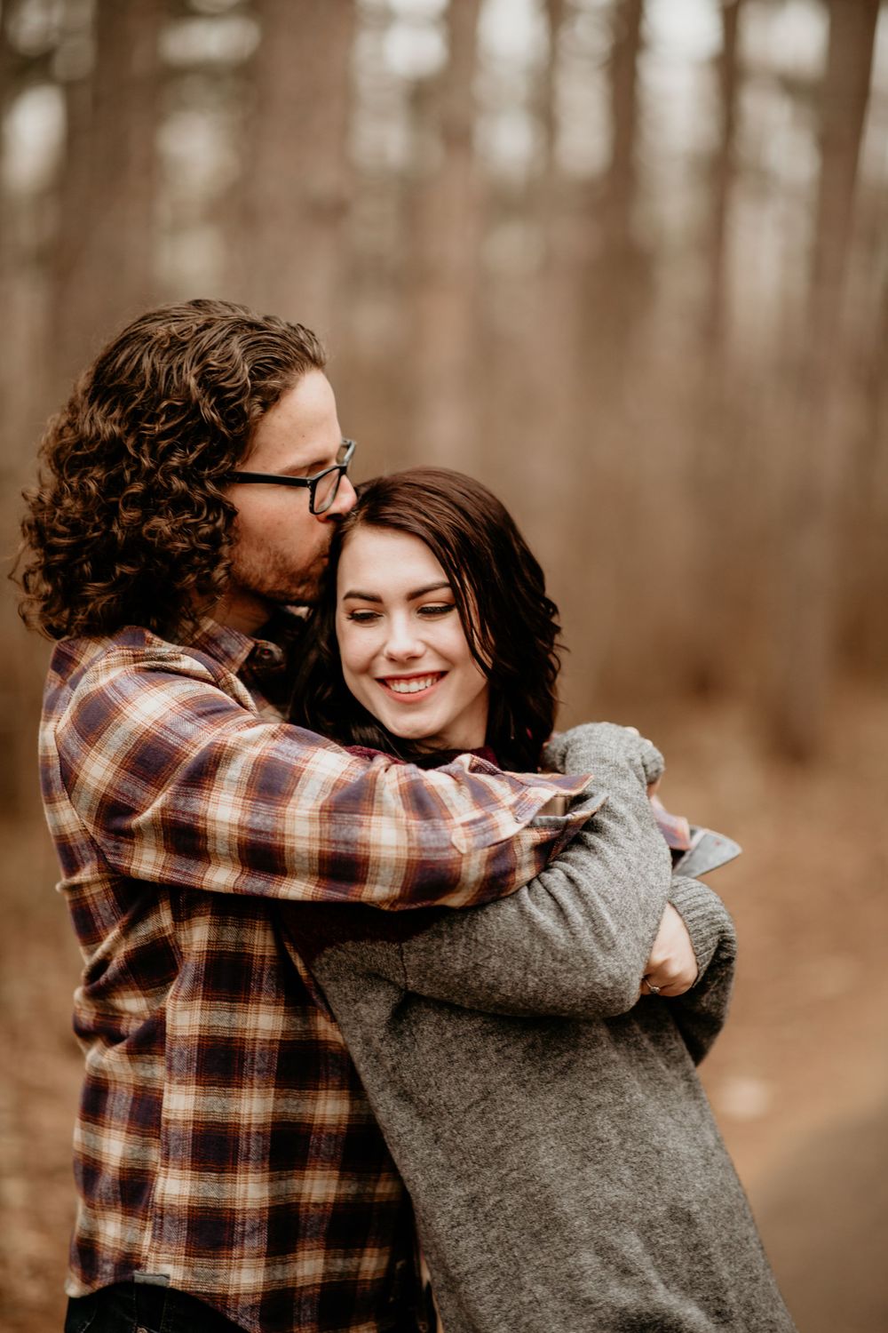 Couple embracing in autumn forest, man kissing woman's temple as she smiles warmly.