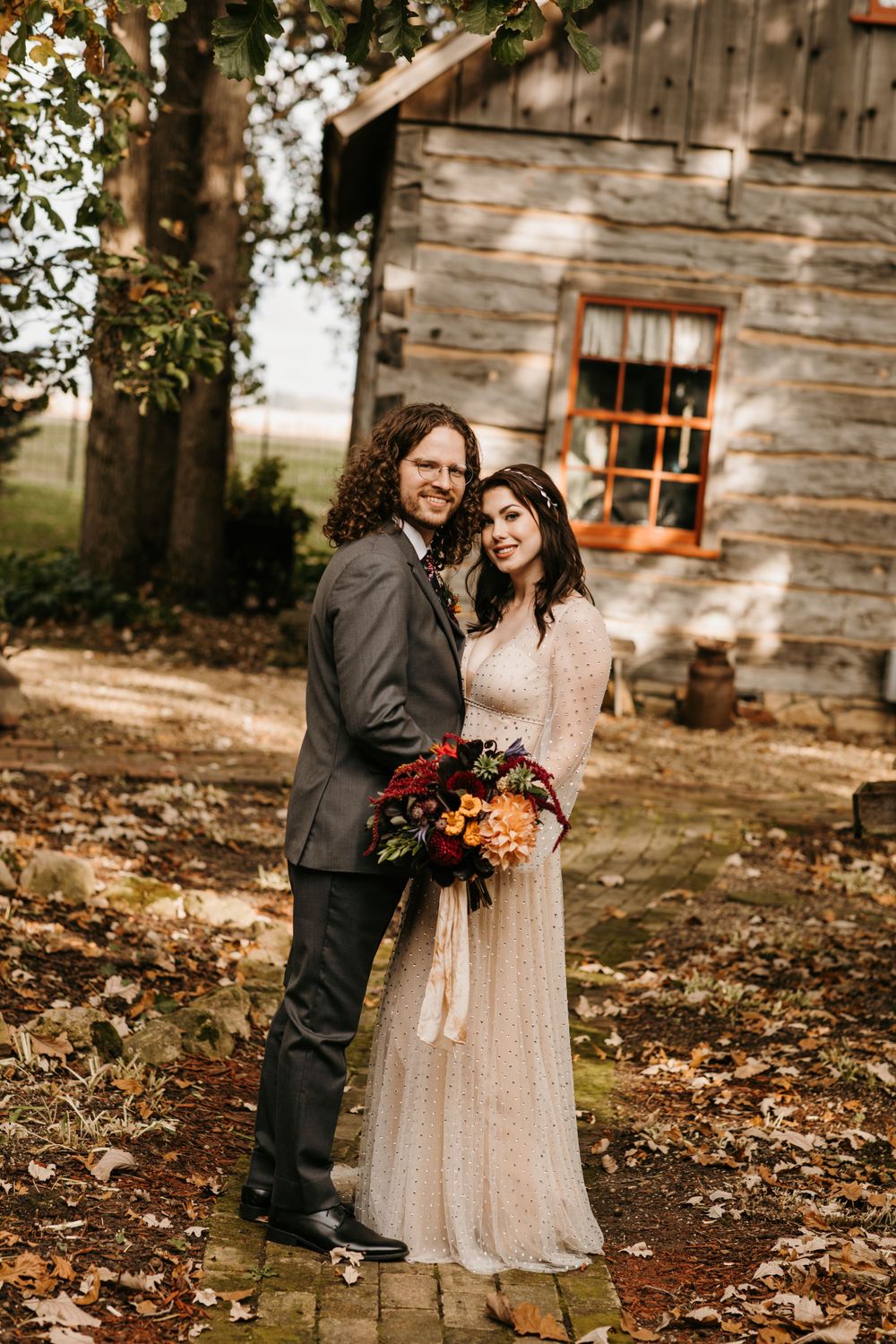 Newlyweds embracing with a colorful fall bouquet in front of a rustic log cabin.