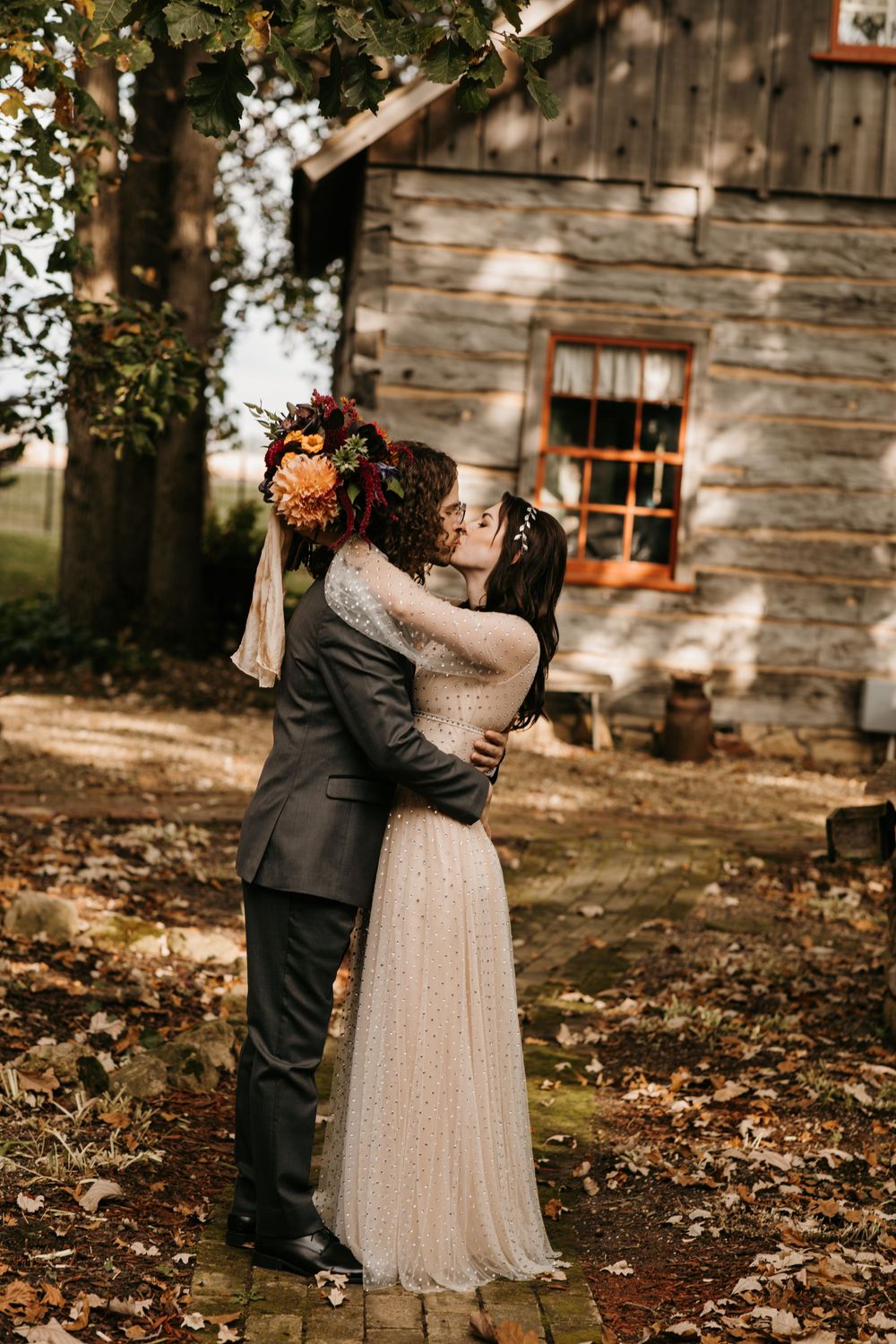 Newlyweds sharing a kiss in front of a rustic log cabin surrounded by fall foliage.