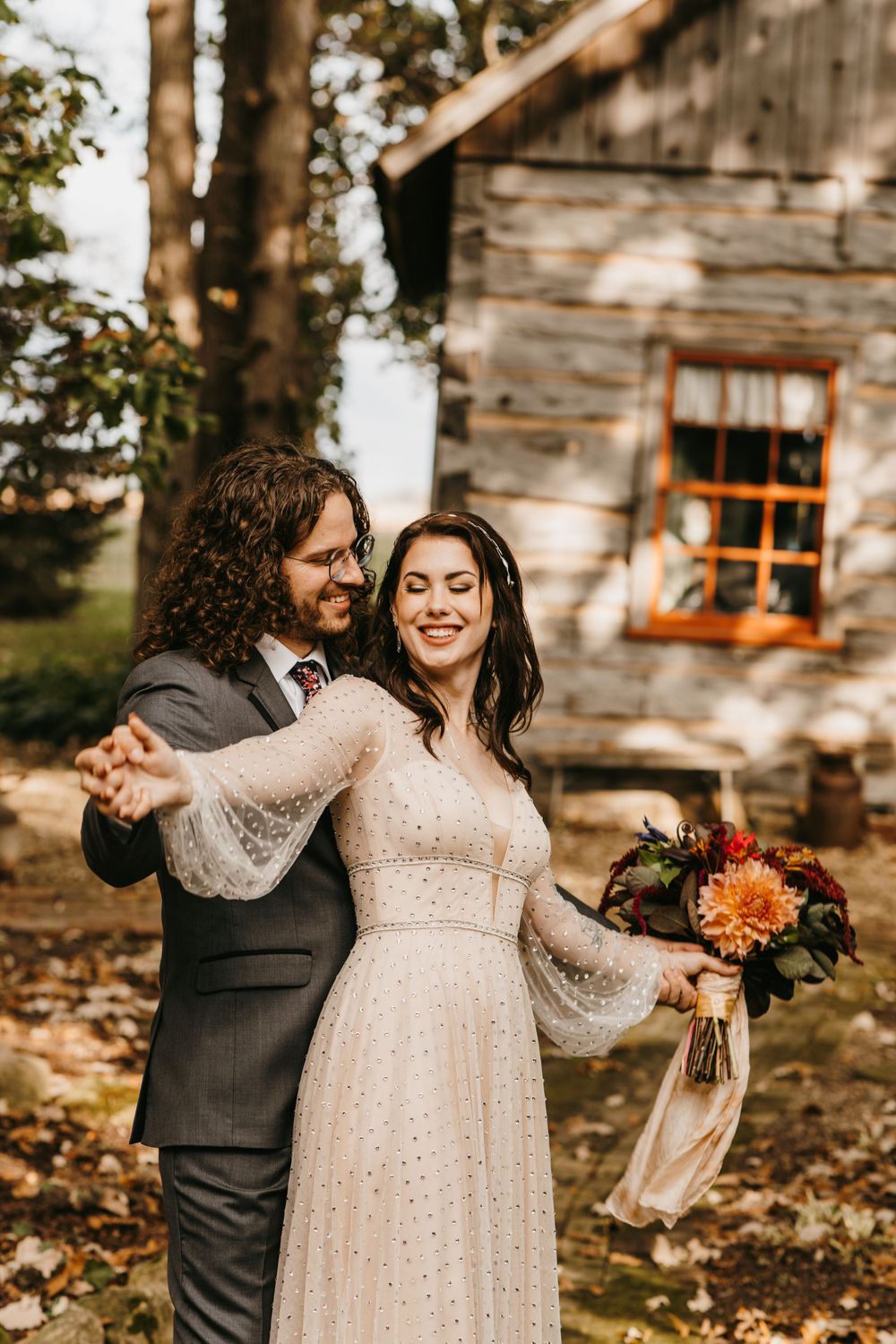 Groom twirling bride in a sparkly gown outside a rustic wooden cabin in autumn.