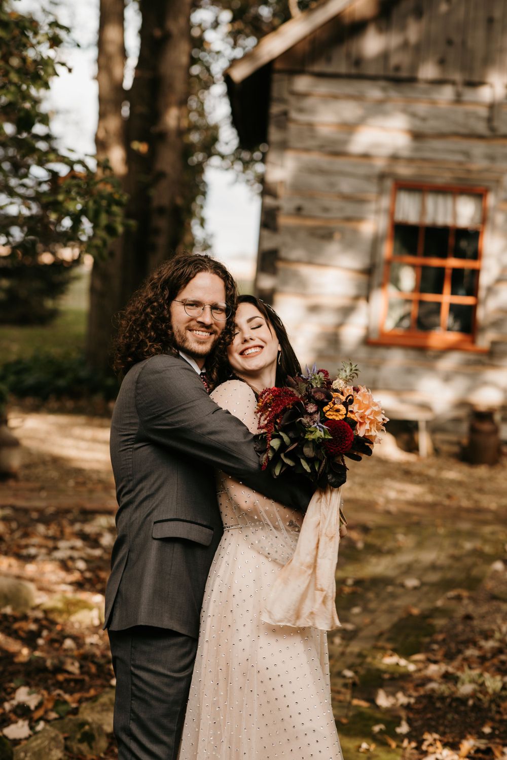Bride laughing joyfully as groom hugs her from behind outside a rustic wooden cabin.