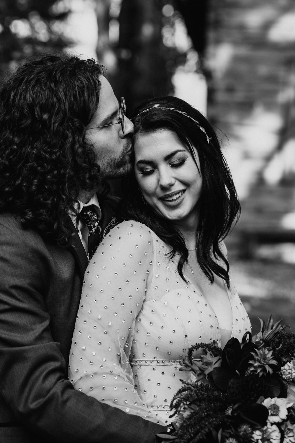 Black and white photo of groom kissing bride on cheek as she smiles holding a bouquet.