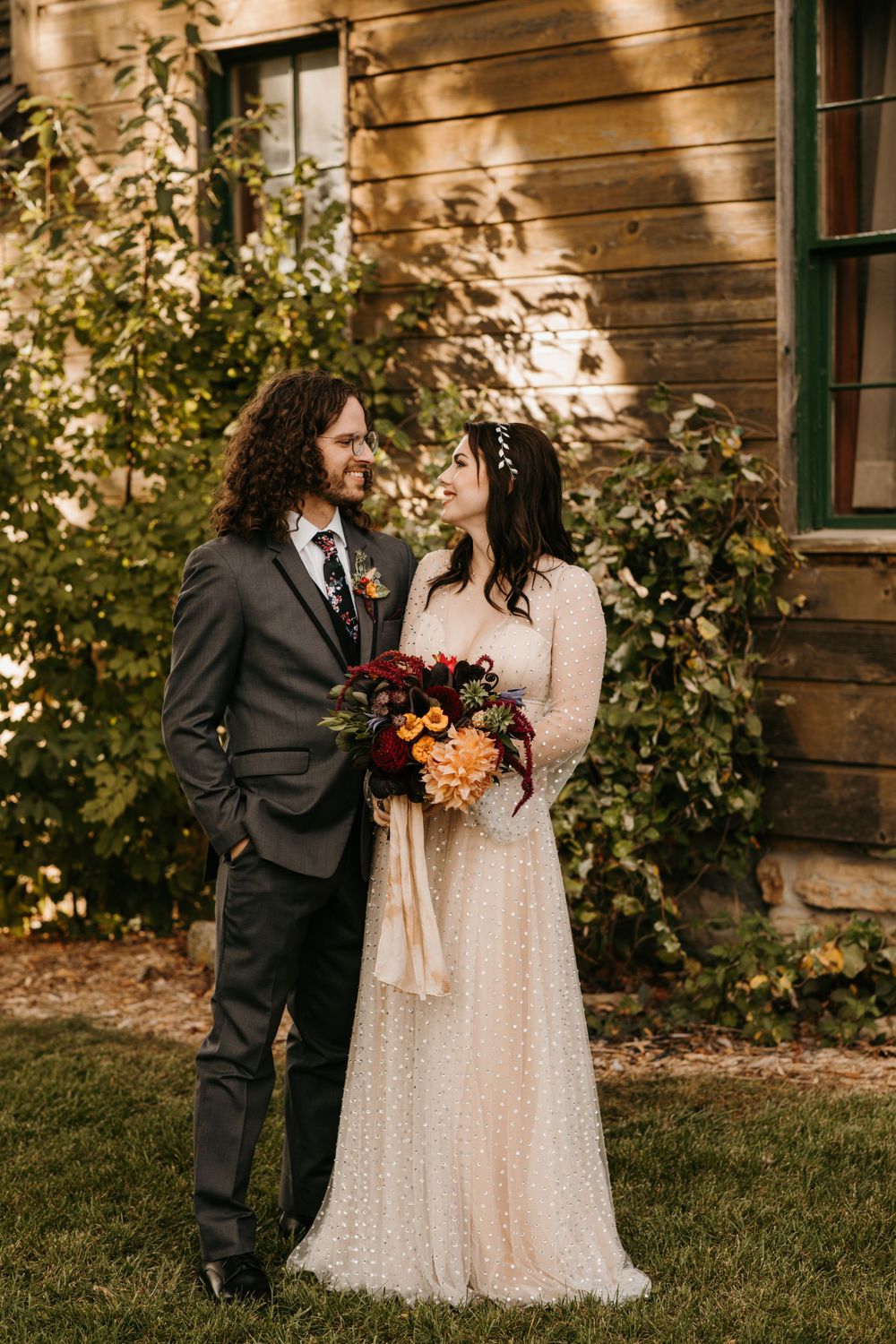Bride and groom gazing at each other by rustic wooden building with fall floral bouquet.