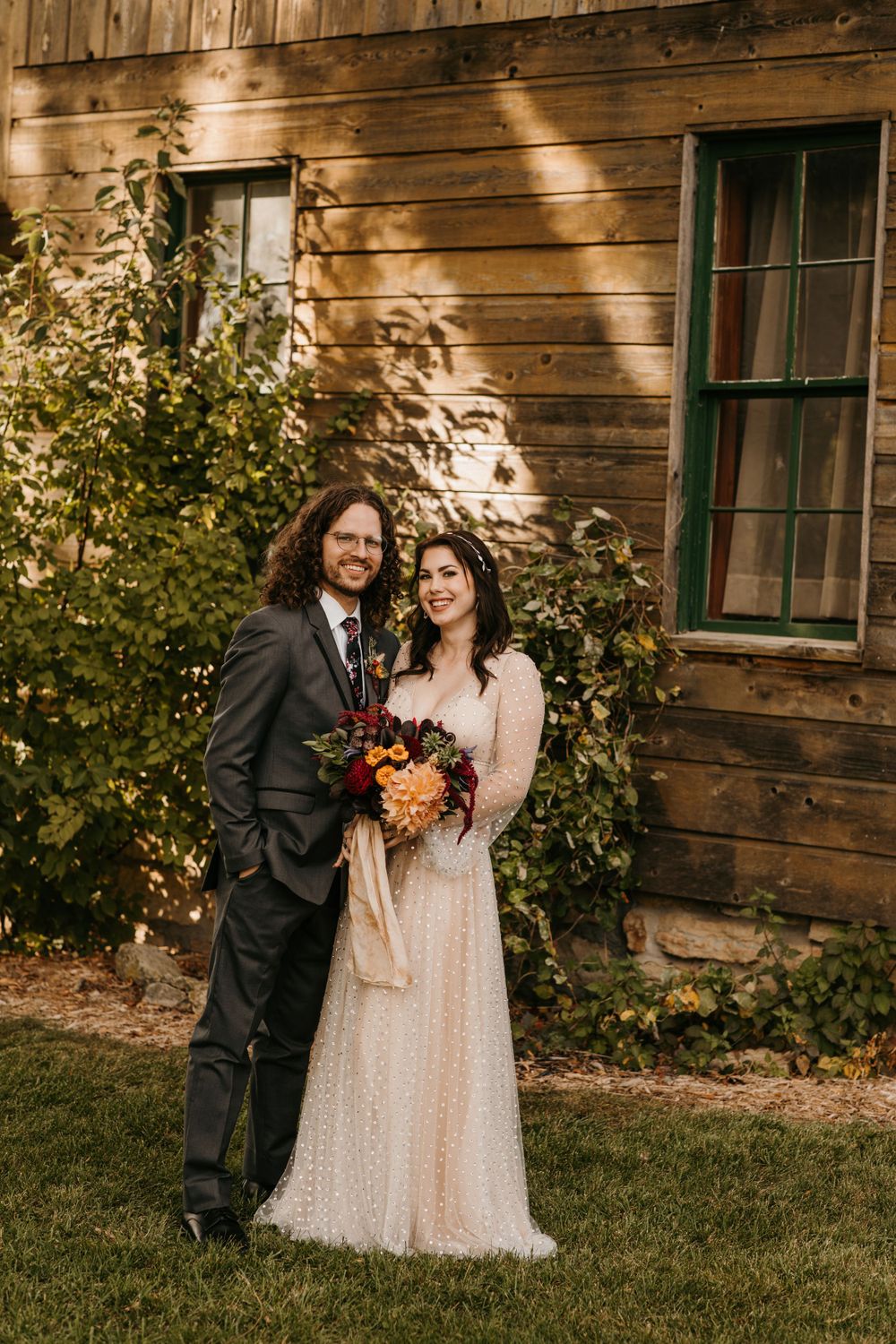 Smiling bride and groom posing together outside rustic wooden barn with autumn bouquet.