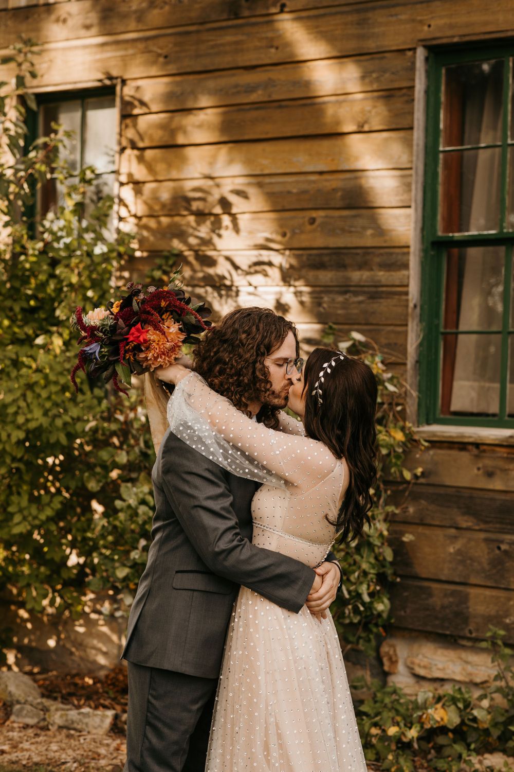 Bride and groom sharing a kiss embracing outside rustic wooden building with fall foliage.