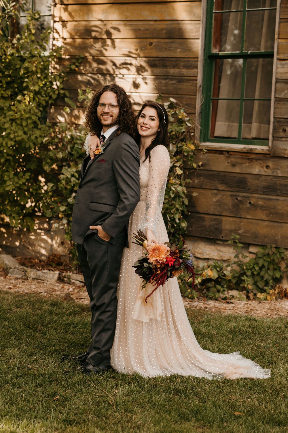 Bride leaning on groom's shoulder smiling outside rustic barn with colorful autumn bouquet.