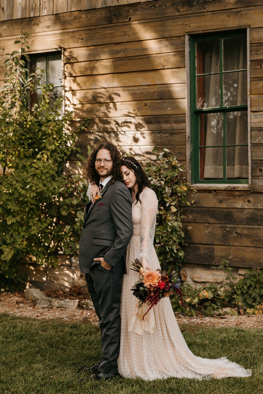 Bride leaning on groom's back outside sunlit rustic barn holding a rich autumn bouquet.