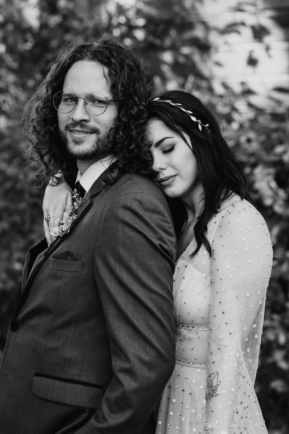 Black and white portrait of bride resting head on groom's shoulder with floral headpiece.