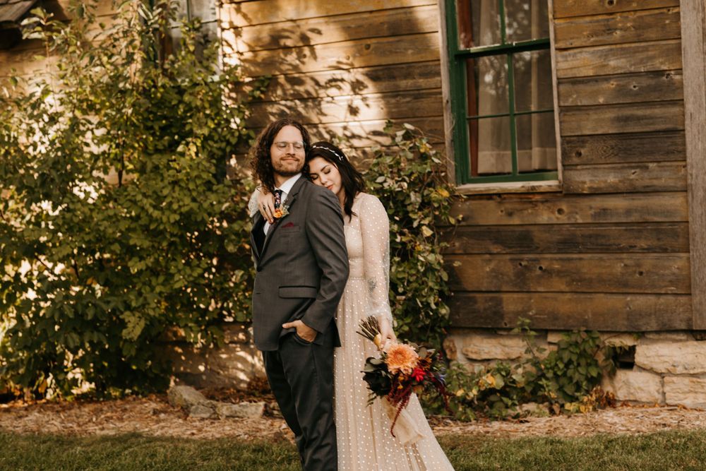 Bride and groom smiling and posing casually outside rustic wooden building with fall bouquet.