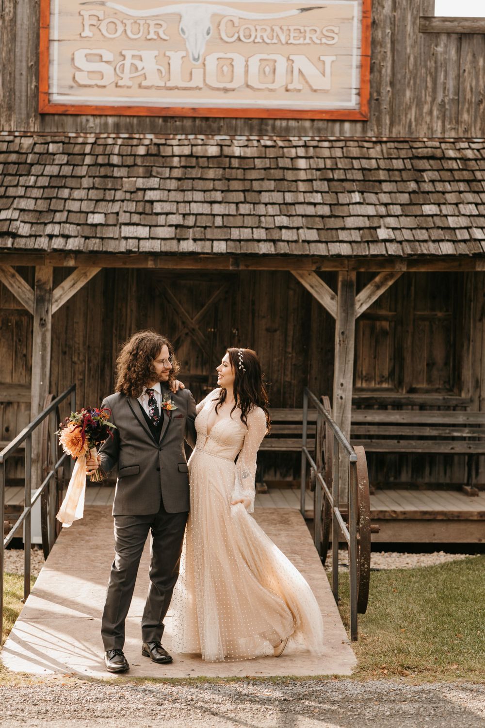 Bride and groom pose outside rustic Four Corners Saloon in wedding attire, bride in flowing champagne gown.