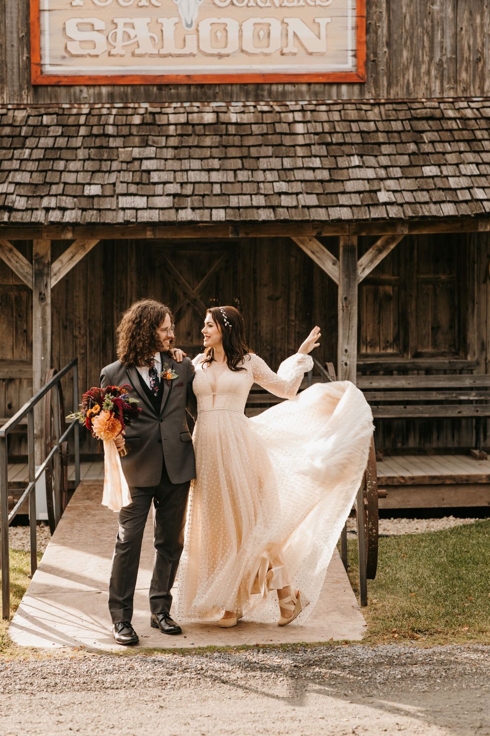 Joyful bride twirls her flowing champagne wedding dress outside a rustic wooden saloon with groom.