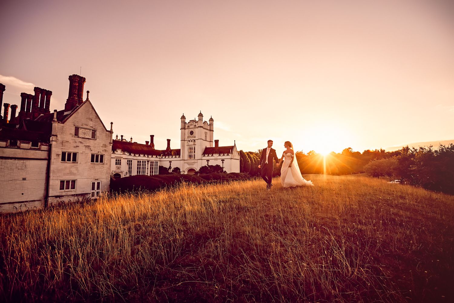 A wedding couple silhouetted against a sunset behind a historic manor house with rustic fields in the foreground.