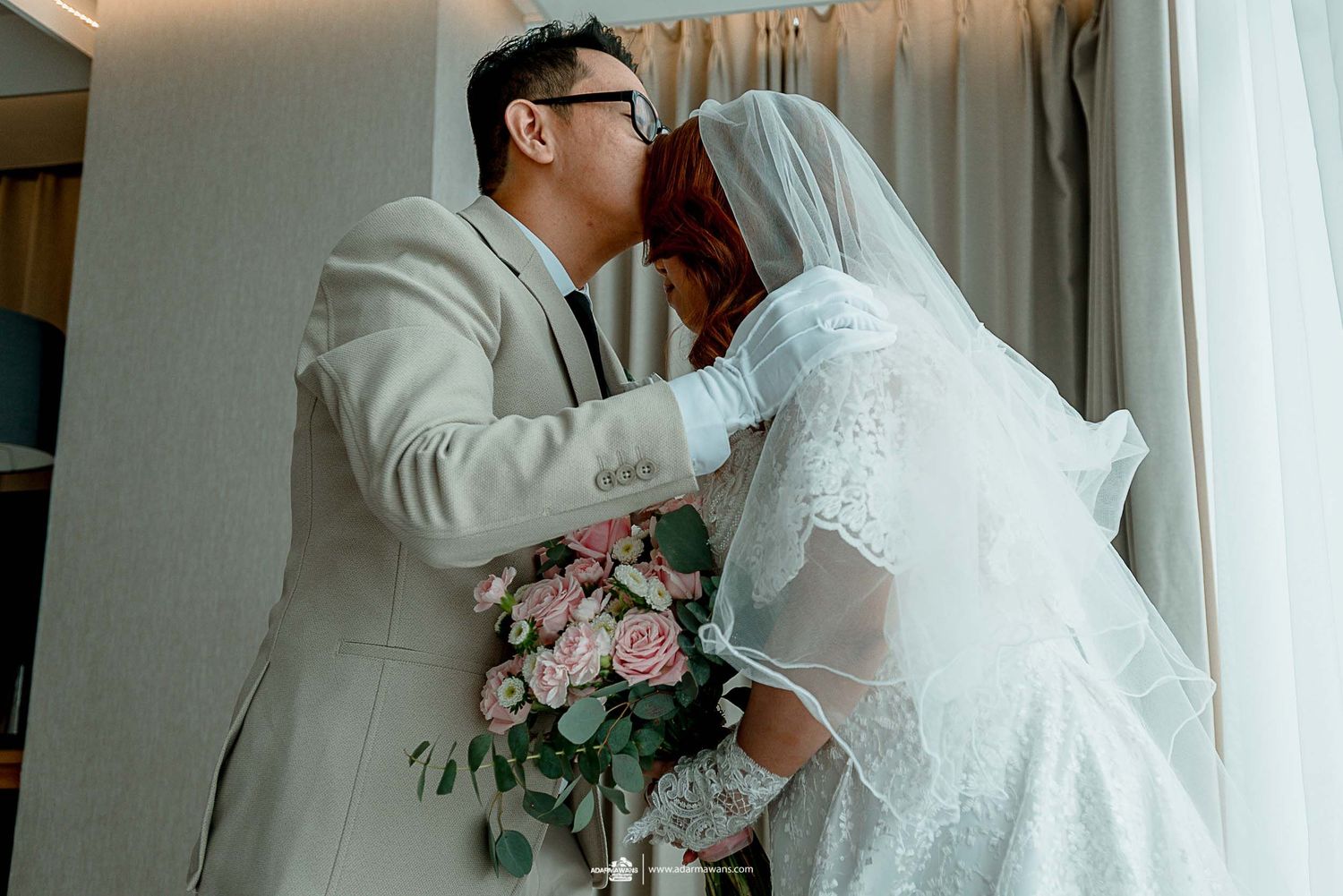 Tender wedding moment with pink floral bouquet in sunlit hotel room.