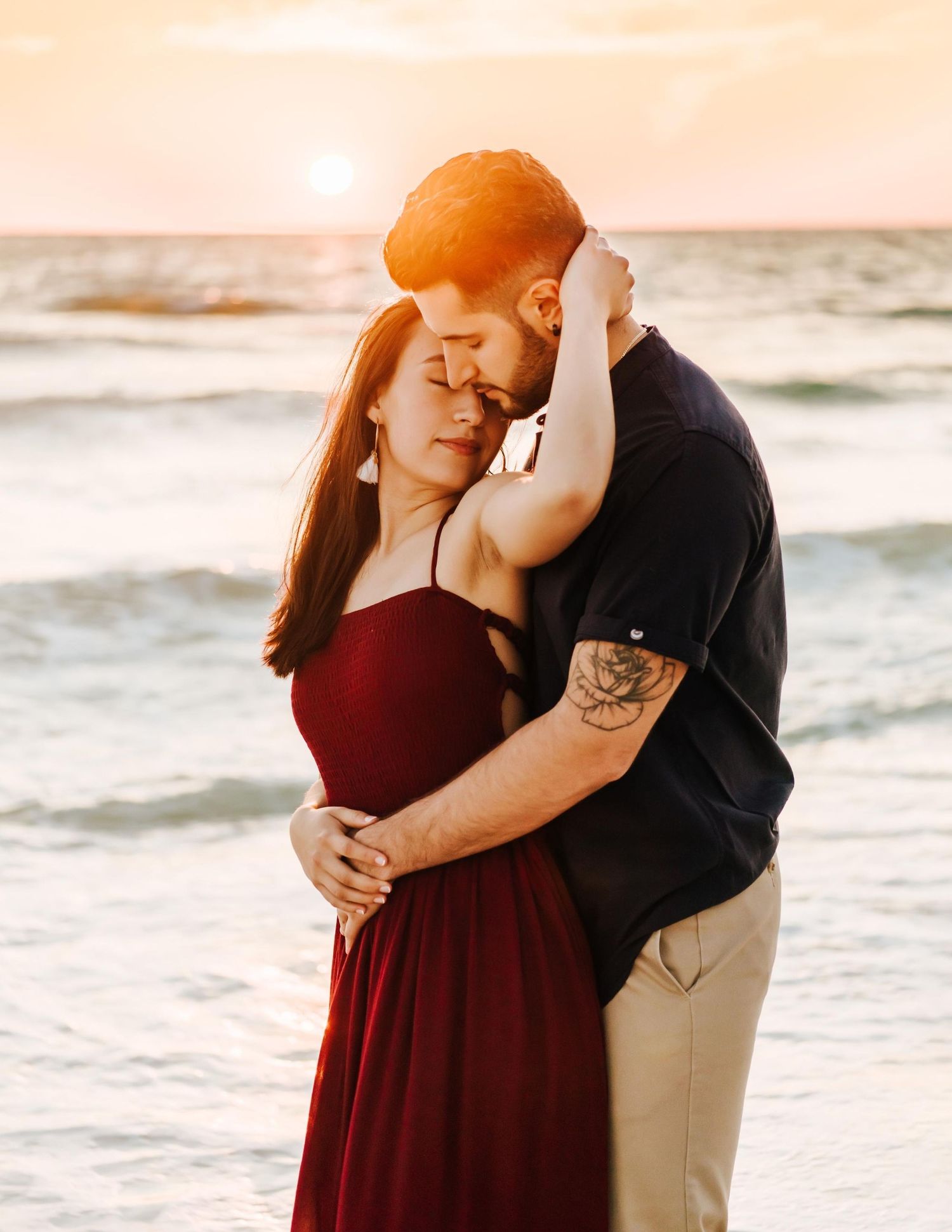 Romantic couple embracing at sunset beach, woman in burgundy dress and man in black shirt with tattoo.