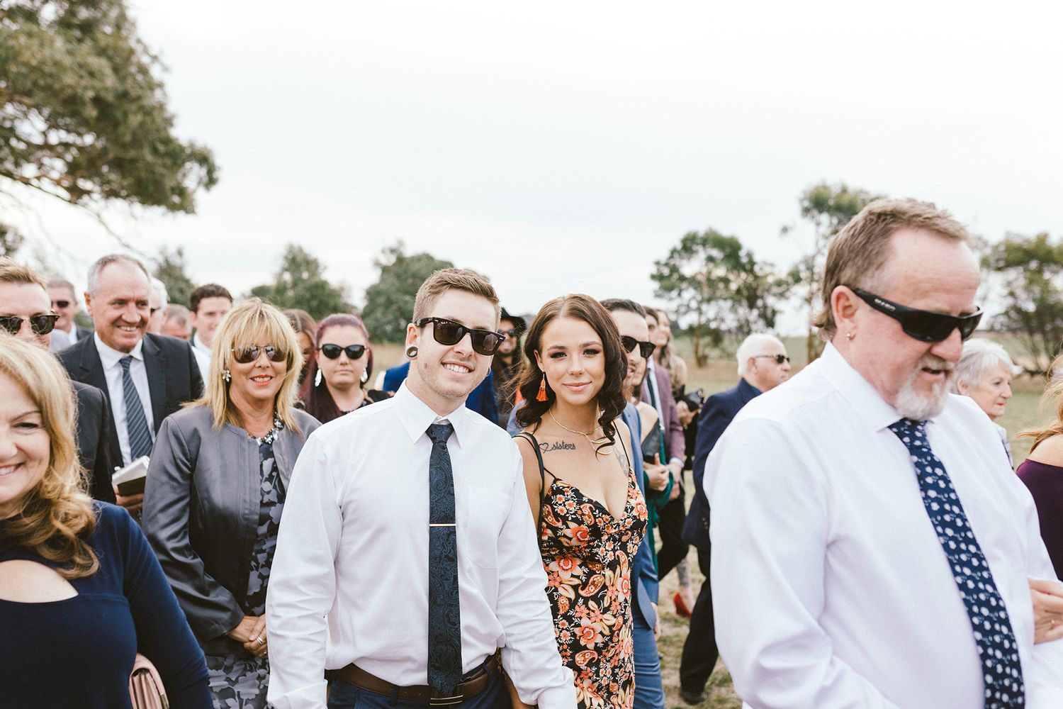 Wedding guests in formal attire gathering outdoors on a sunny day.
