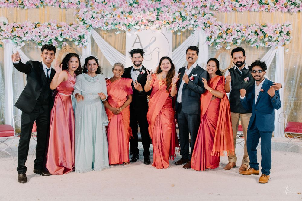 Wedding party group photo with people wearing coral and grey formal attire against a floral backdrop.