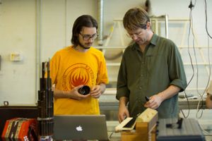 Two people collaborating on a project with laptop and equipment in workshop setting.