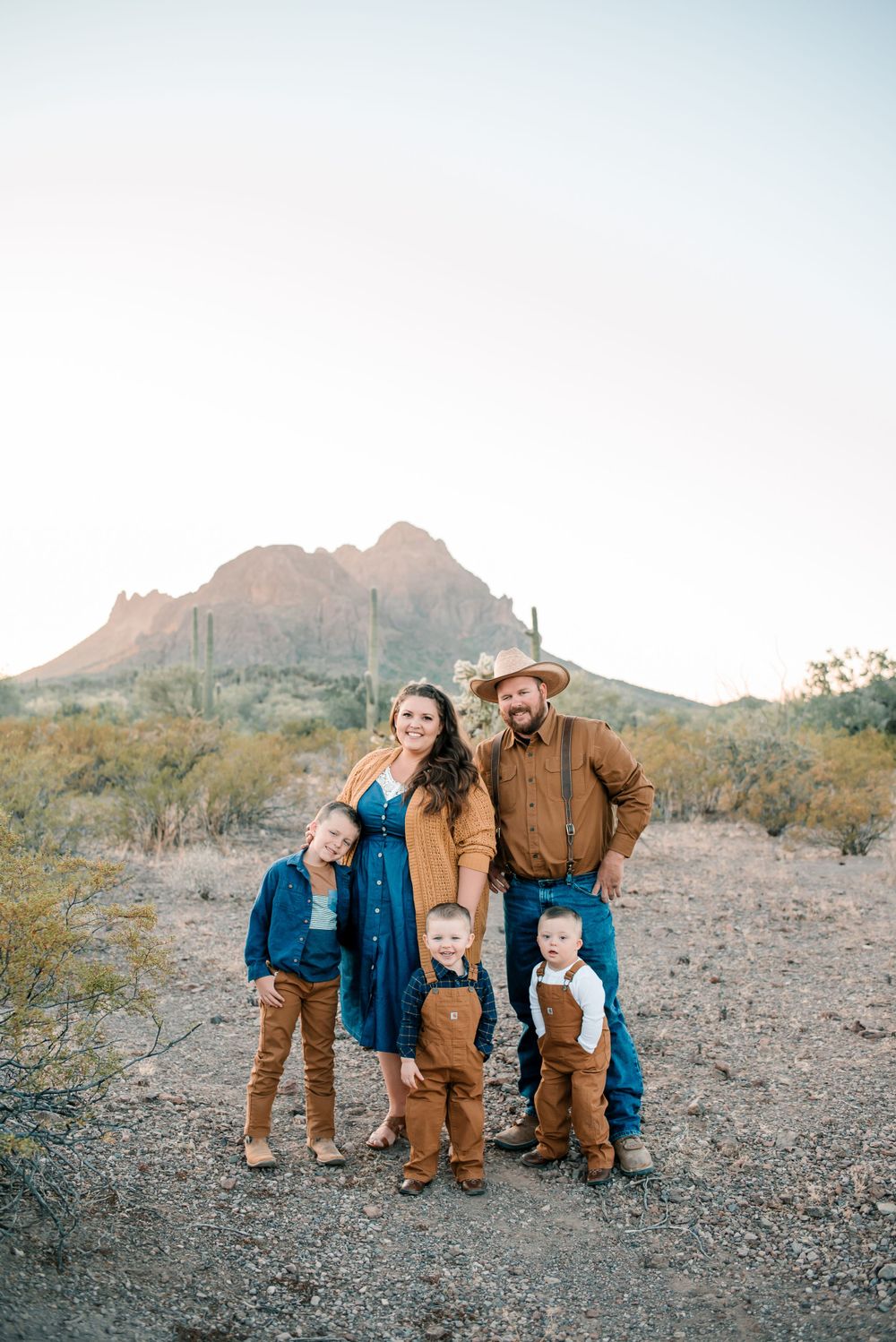Family wearing coordinated blue and brown outfits poses together in desert landscape with mountains in background.