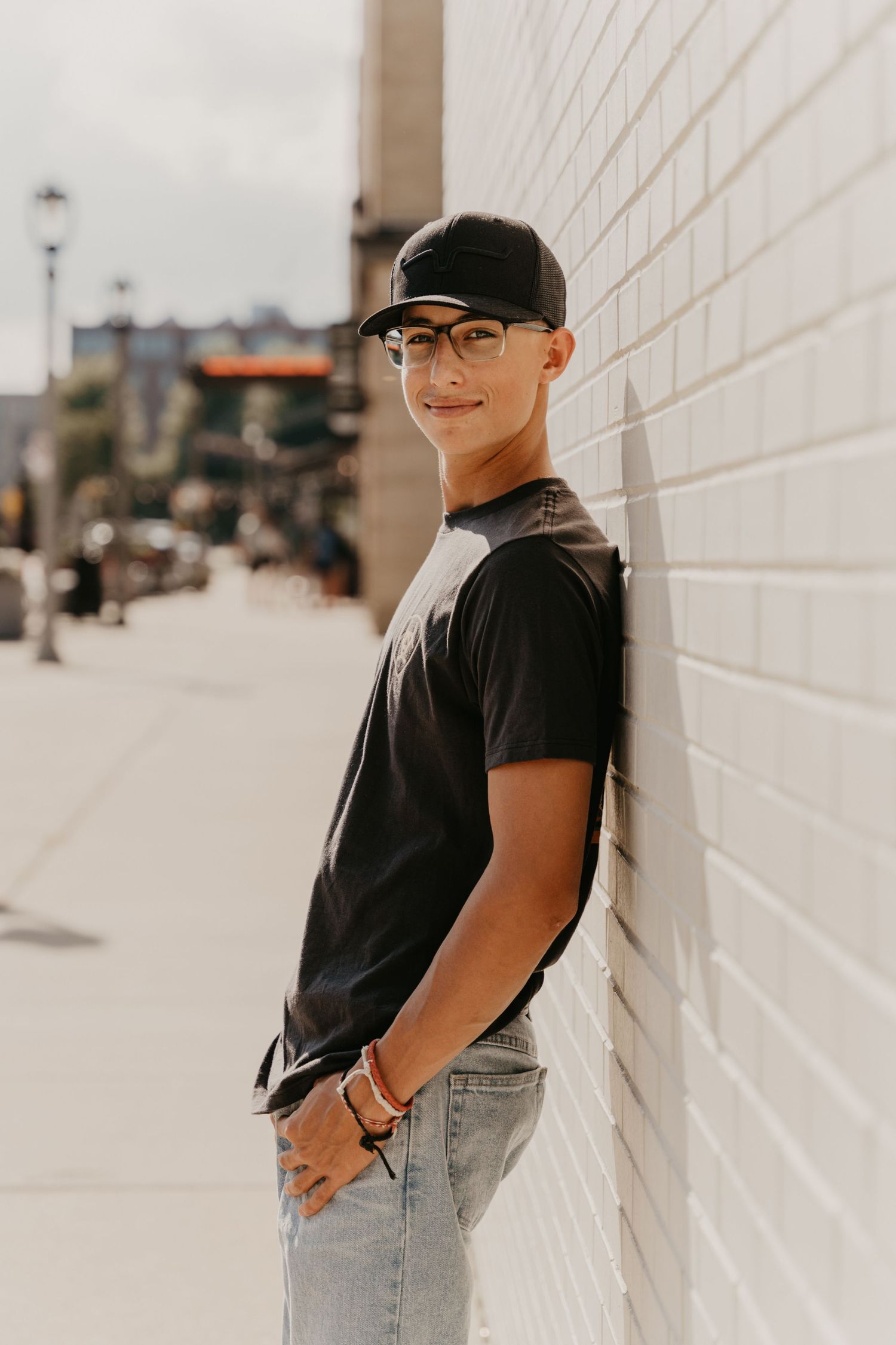 Casual portrait of someone in baseball cap leaning against white wall on sunny urban street.