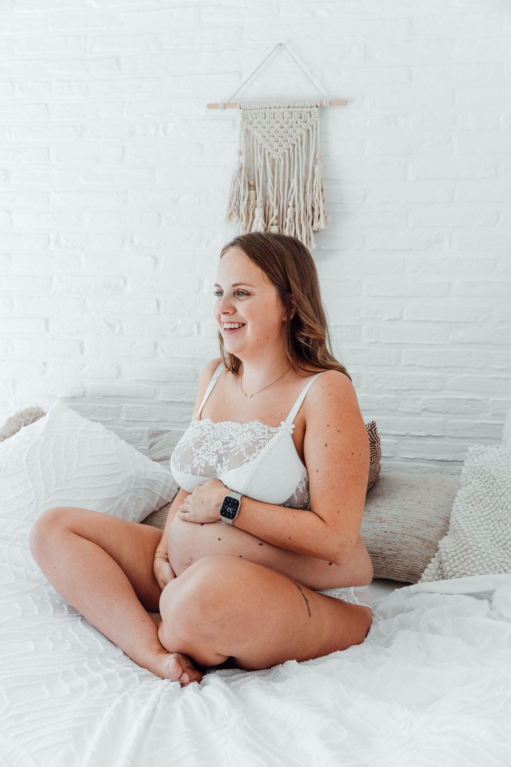 Pregnant woman in white loungewear sits comfortably on bed with macrame wall hanging in background.