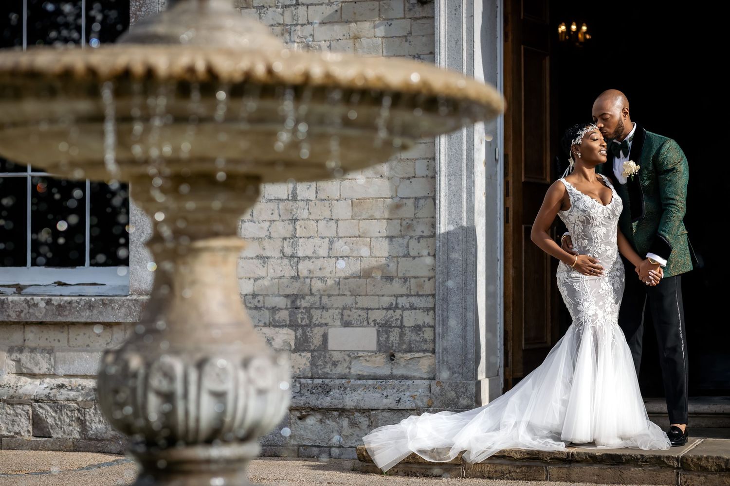 Romantic couple poses beside ornate stone fountain during elegant wedding photoshoot.