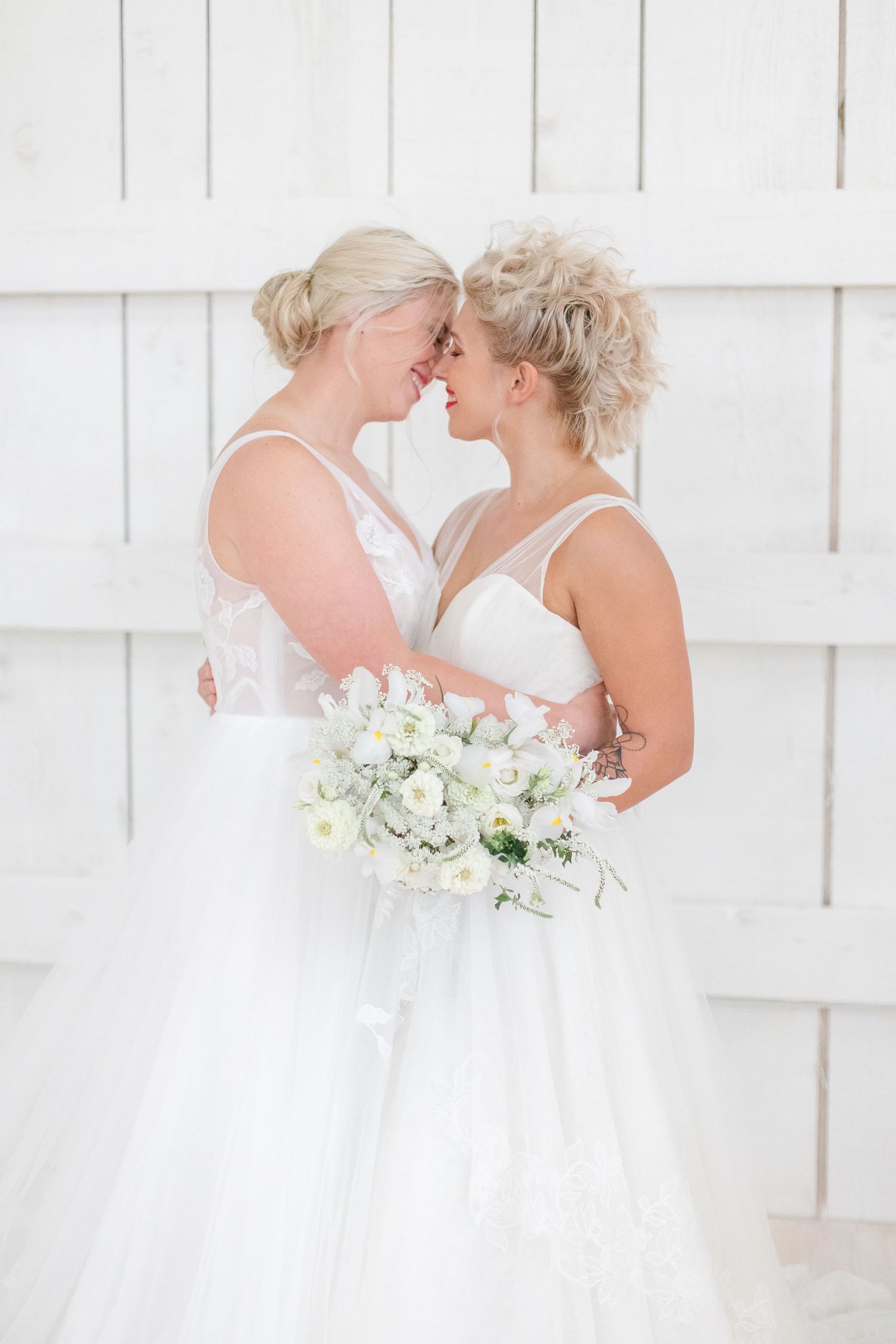 Couple in matching white wedding dresses embracing against a white shiplap wall.