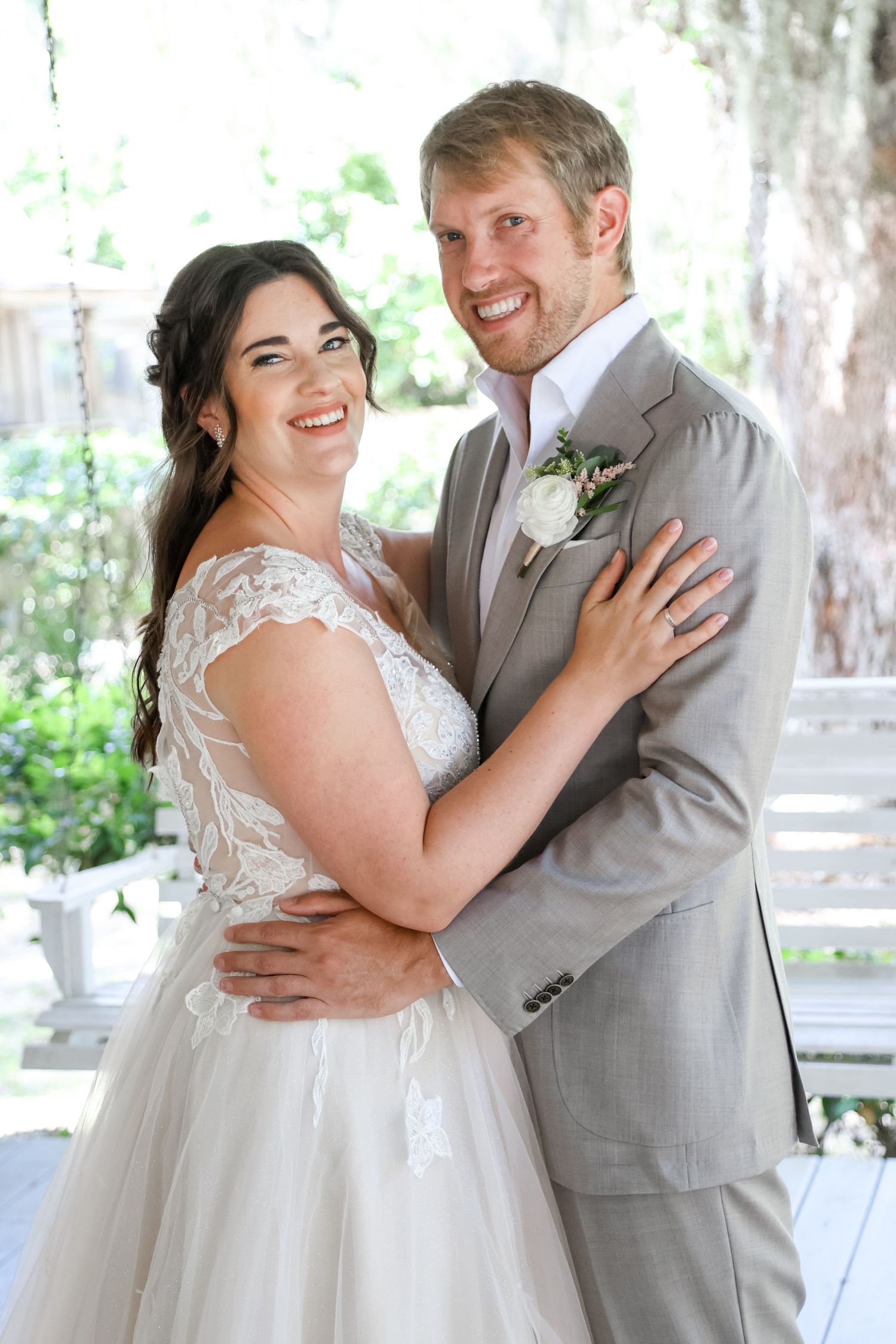 A happy couple embraces on their wedding day, wearing a white lace gown and light gray suit in an outdoor setting.