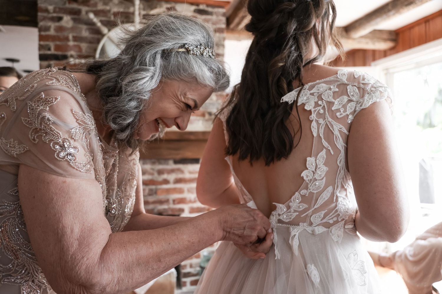 A tender moment of helping button up a delicate white lace wedding dress against exposed brick walls.