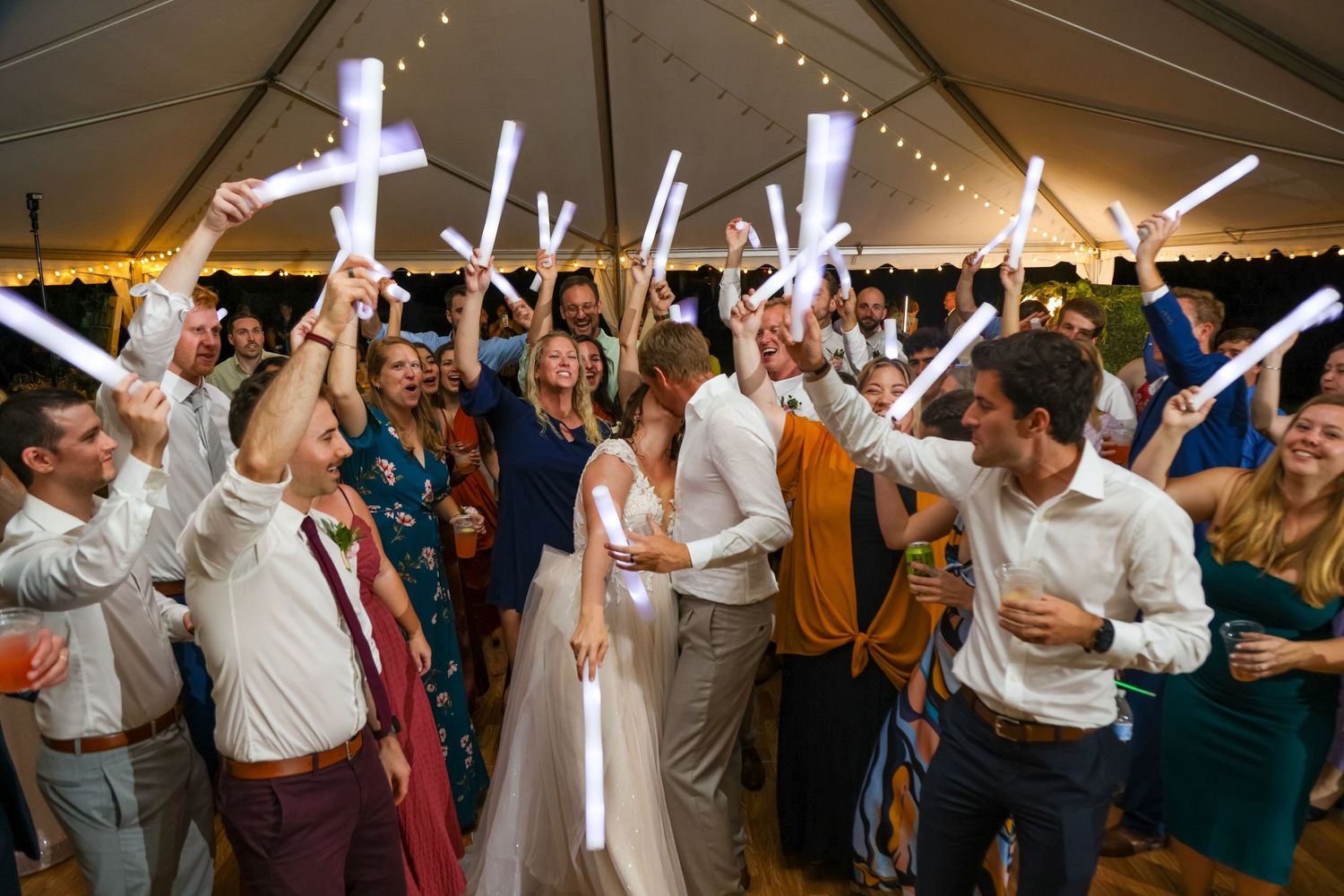 Wedding reception dance party under tent with crowd holding illuminated LED foam glow sticks in celebration.