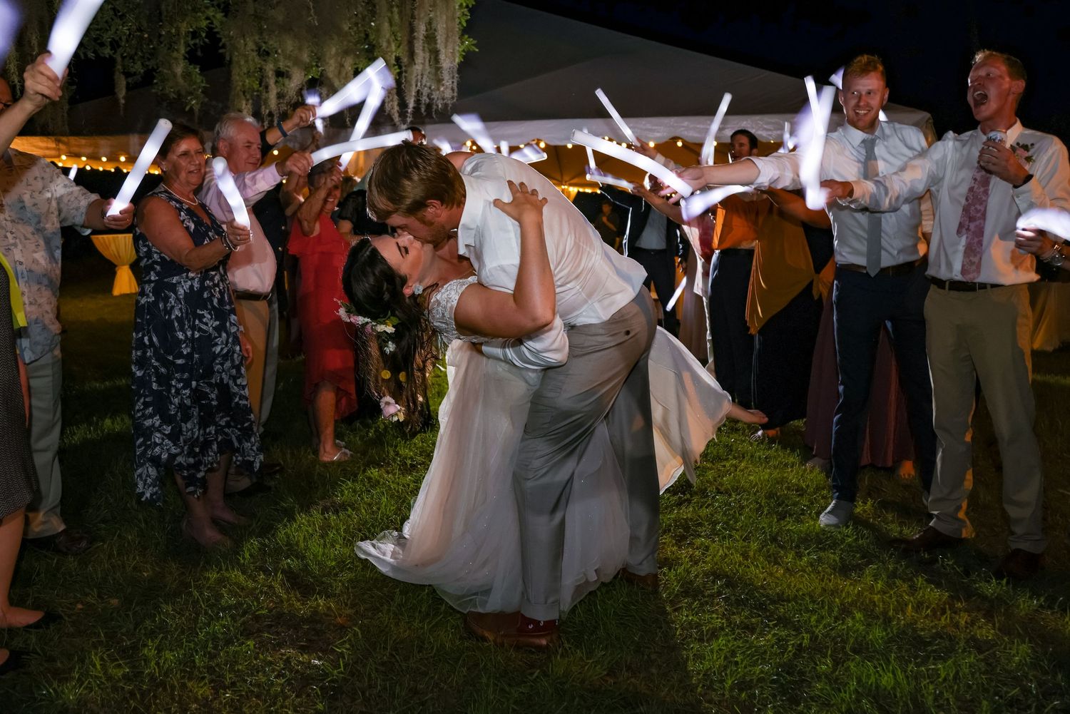 Wedding attendees hold glowing light sticks while dancing outdoors at night.