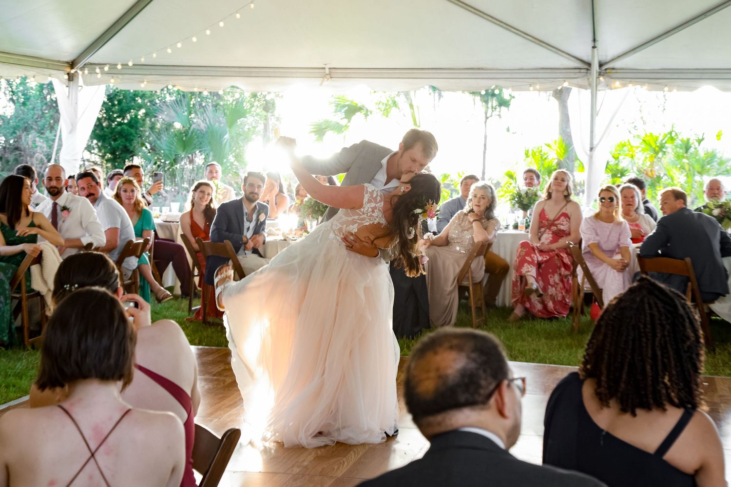A couple shares their first dance under a white tent as wedding guests watch and celebrate.