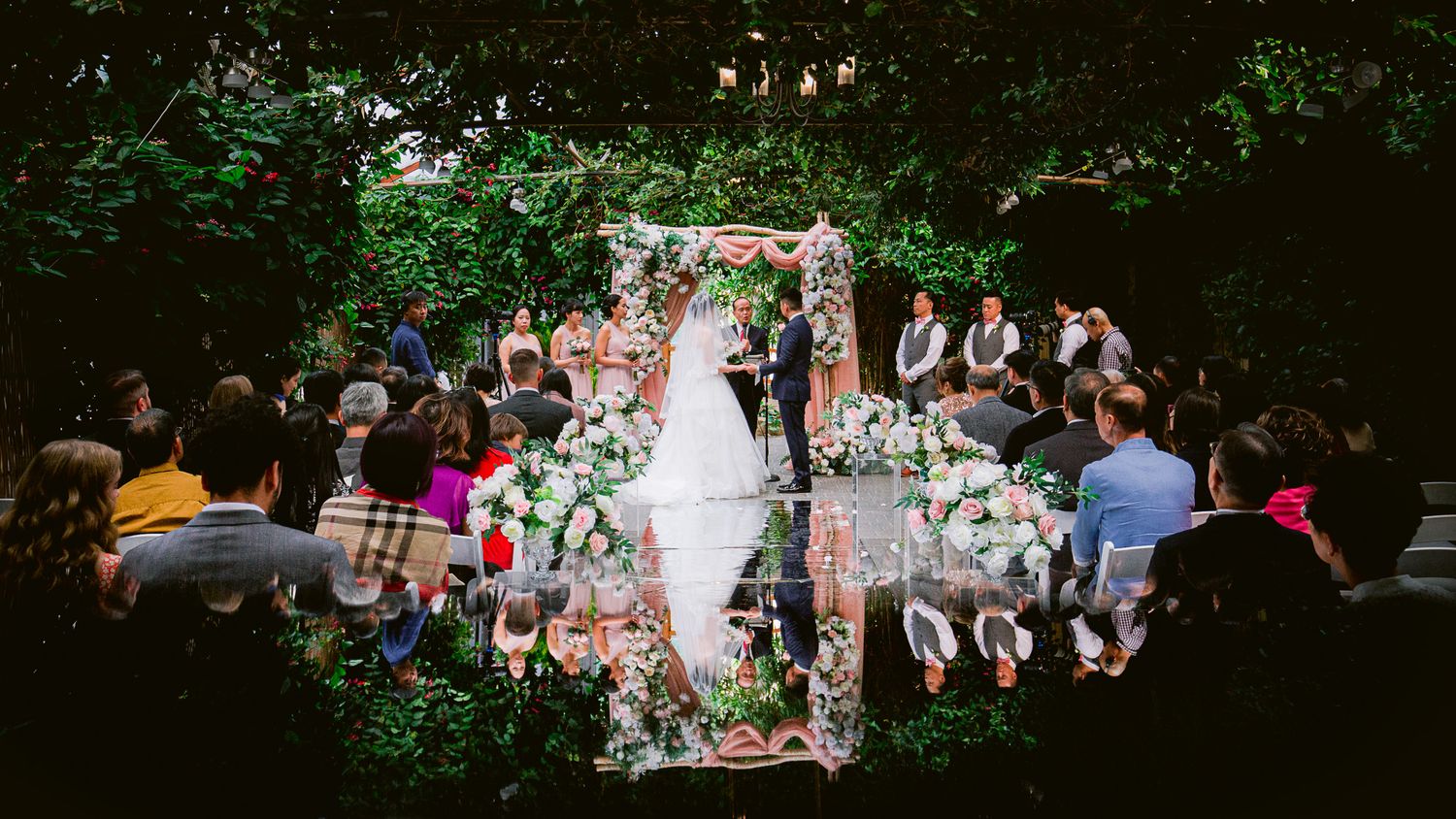 Outdoor wedding ceremony in a lush garden setting with floral arch and gathered guests on both sides.