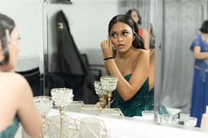 Person in emerald green dress applies makeup at vanity table with decorative crystal glasses.