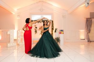 Two people in contrasting red and emerald gowns pose playfully in elegant venue.