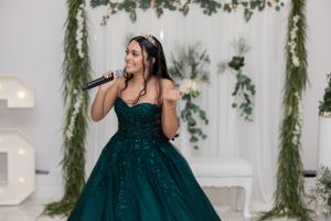 A performer in a dark green sequined ballgown sings into a microphone against a decorated backdrop.