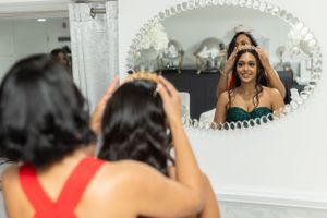 Person in red dress helping another in formal attire style hair in ornate oval mirror.