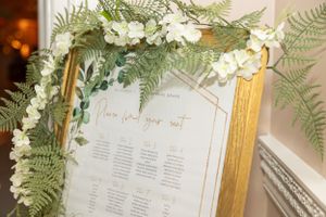 Gold-framed seating chart decorated with white flowers and ferns.