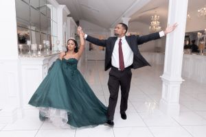 Couple in emerald gown and suit pose dramatically in elegant white ballroom.