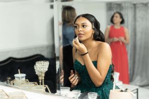 Person in sparkly green gown touching up makeup at elegant dressing table with crystal glasses.