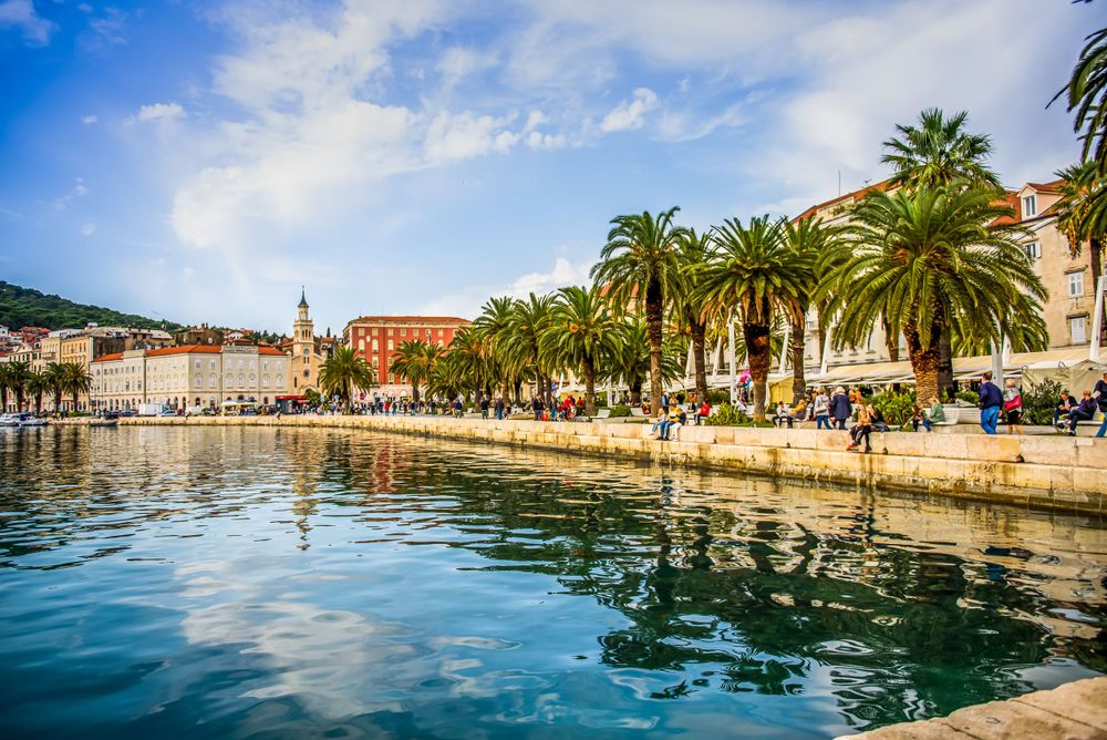 Palm tree lined waterfront promenade in Split, Croatia with historic buildings reflected in calm Mediterranean waters.