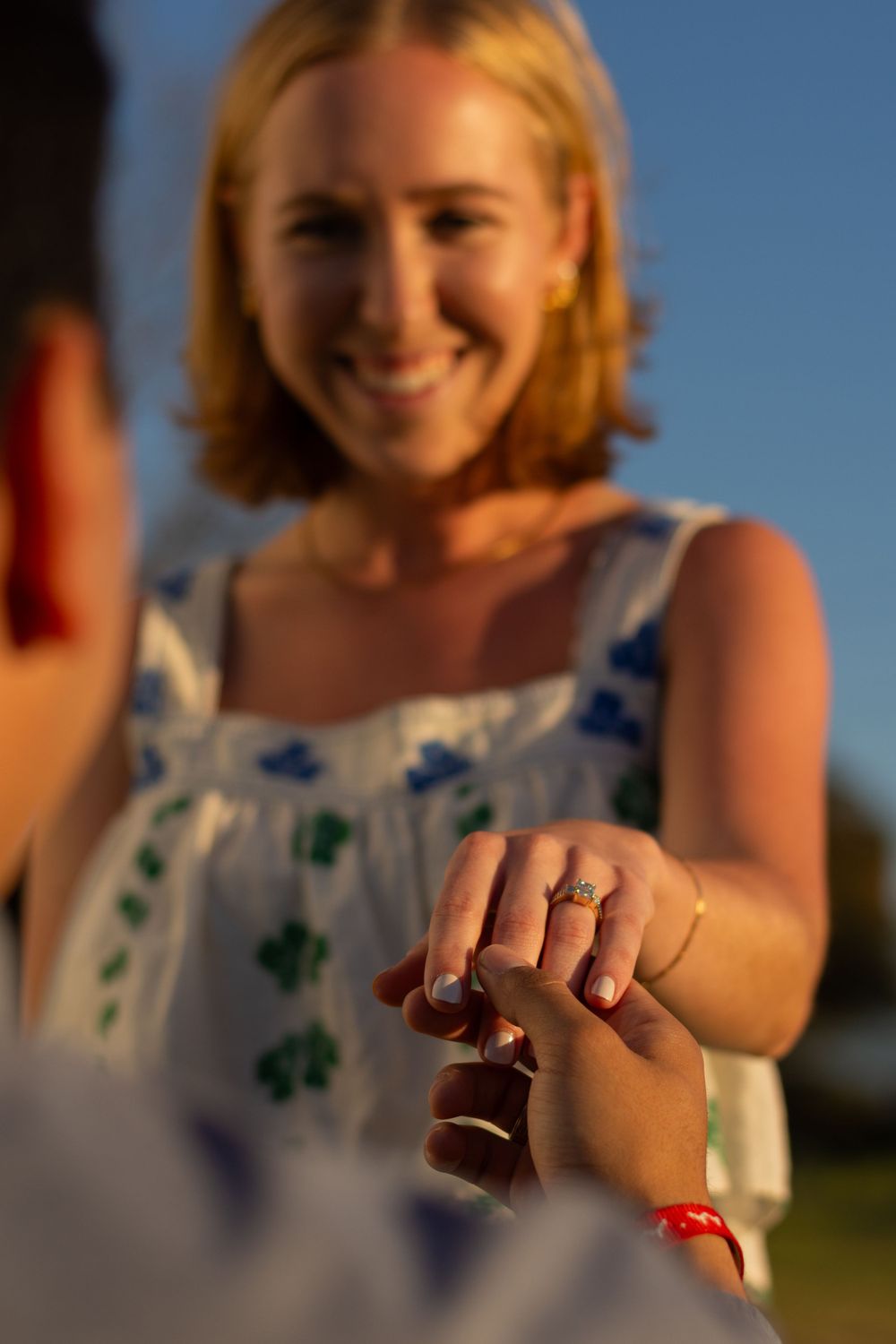 Over the shoulder of a man holding his new fiancée's ring hand as she smiles at him during a sunset couple photo session in Forth Worth.