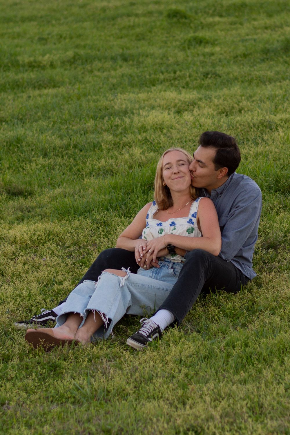 A man kisses his fiancée's cheek as they sit in a grass field in a Fort Worth park at sunset. She closes her eyes as she feels loved by him.