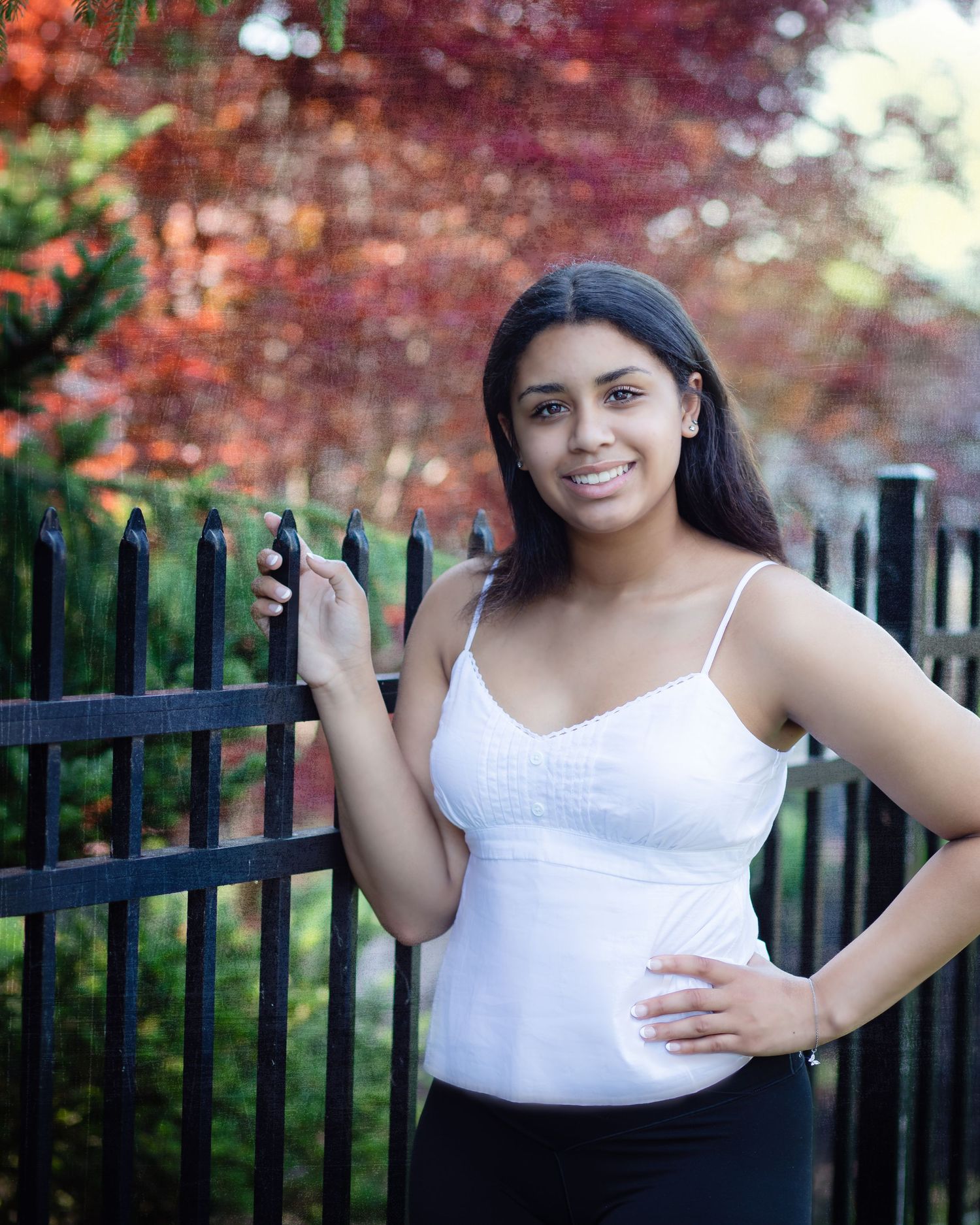 A person in a white top stands by a black iron fence with red autumn foliage in the background.