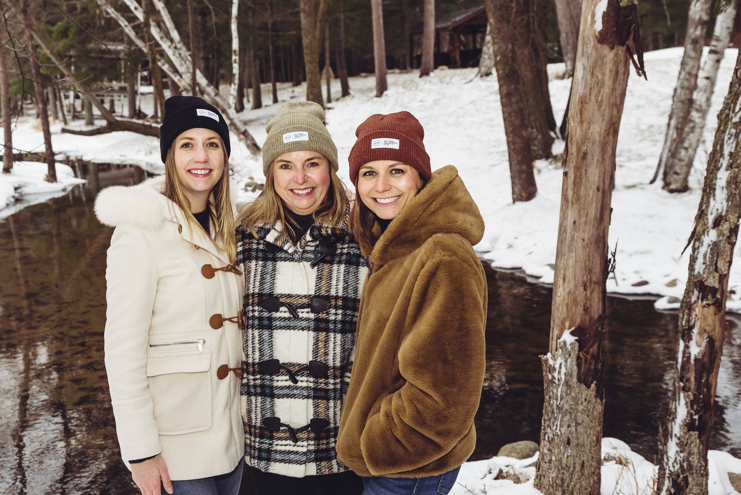 Three friends in winter coats stand together in snowy woods near a stream.