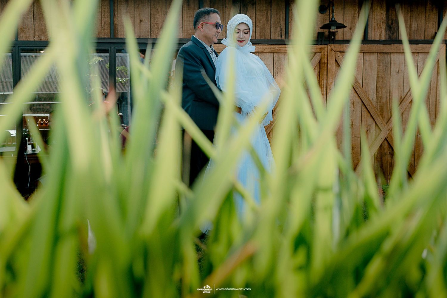 Couple walking together through tall grass with rustic barn in background.