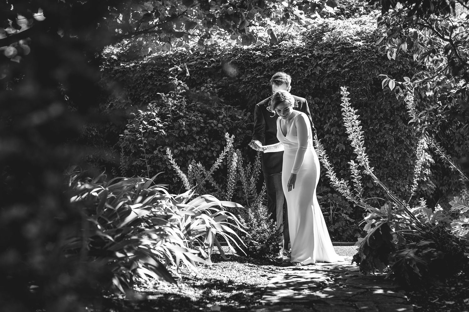 Black and white photo of an elegant wedding dress silhouetted against a garden path surrounded by lush foliage.